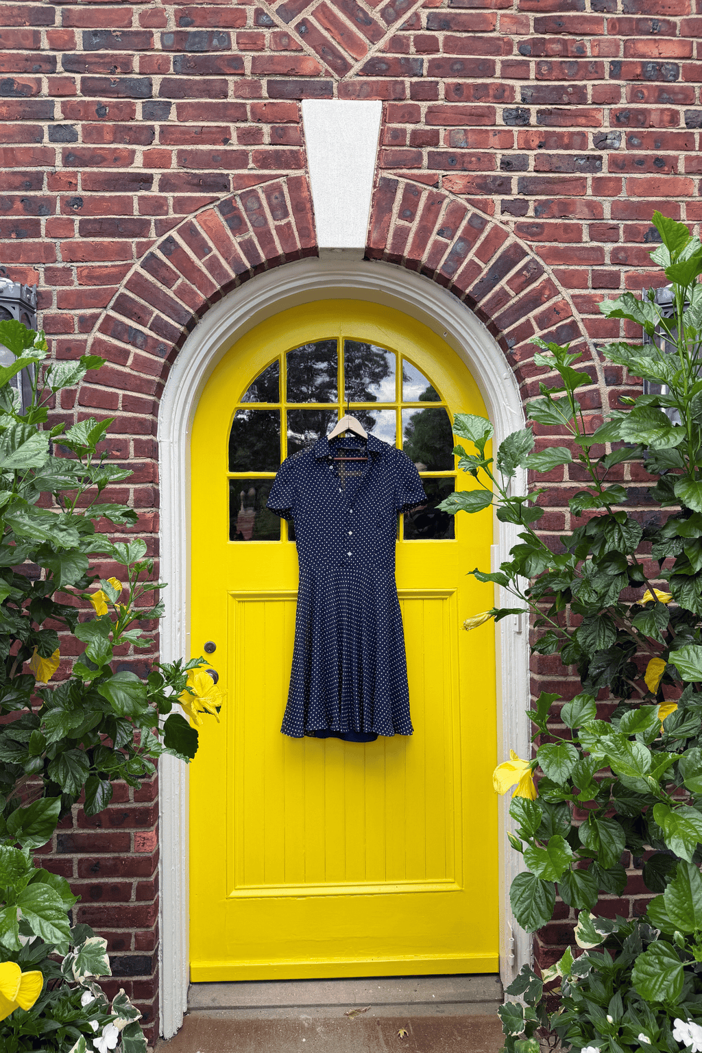 Polo Ralph Lauren navy silk georgette polka dot shirt dress with short sleeves and pleated skirt in front of a yellow door. 