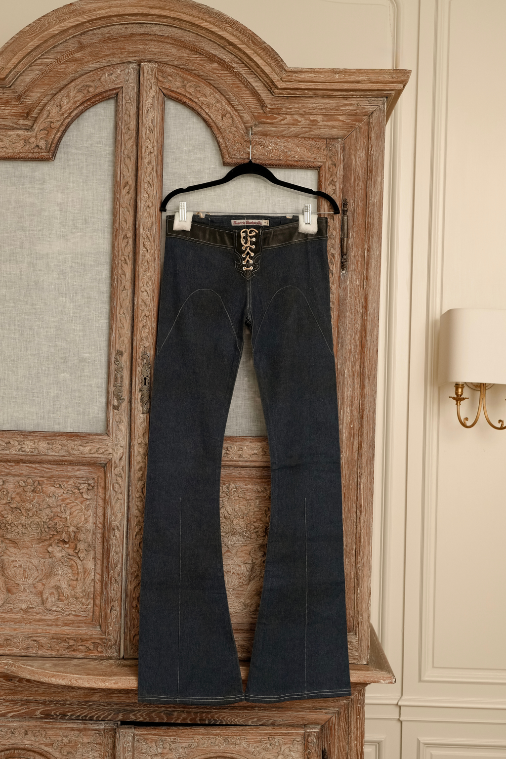 Front view of dark wash Electric Barbarella  jeans hanging on a wooden cabinet, featuring a black leather-trimmed waistband and a white lace-up leather closure at the fly.