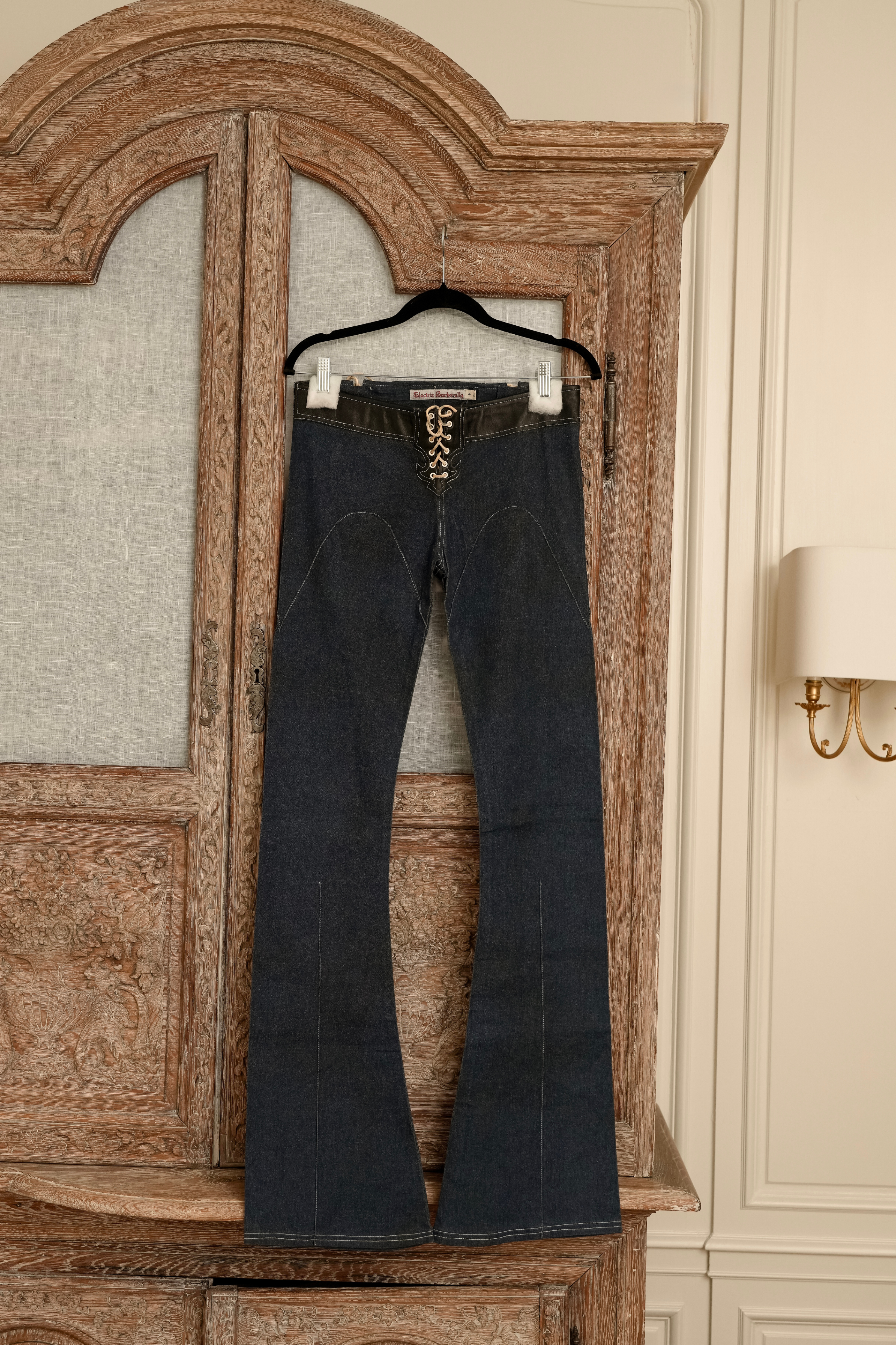 Front view of dark wash Electric Barbarella  jeans hanging on a wooden cabinet, featuring a black leather-trimmed waistband and a white lace-up leather closure at the fly.