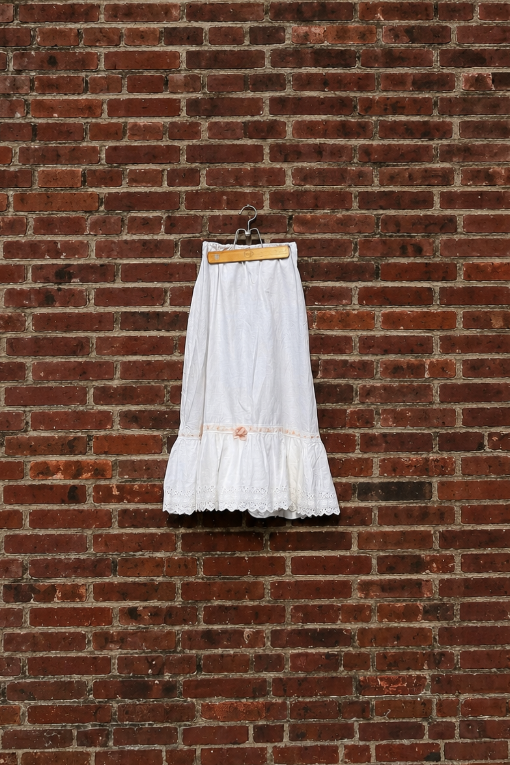 A white antique edwardian skirt with a lace hem hangs from a vintage wooden hanger against a rednbrick wall.