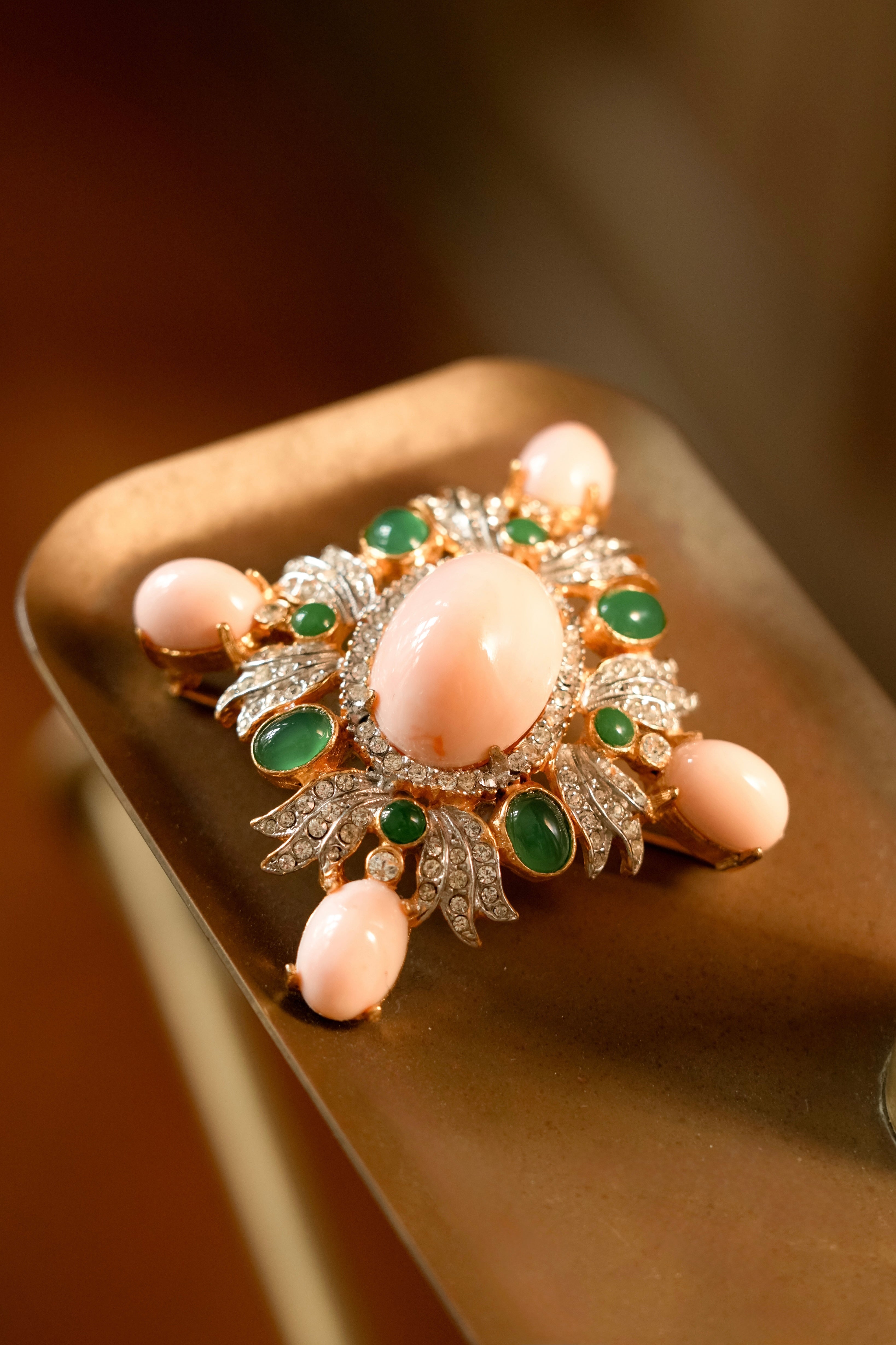 Close-up of a vintage Pauline Rader Maltese cross style brooch featuring a large center pink coral cabochon, surrounded by green jade-colored stones, rhinestones, and four outer coral stones set in textured gold metal.