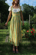 A woman wearing a prairie dress,  with a green, yellow, and white floral pattern, a white ruffled bodice, and puffed sleeves, standing in a garden setting.