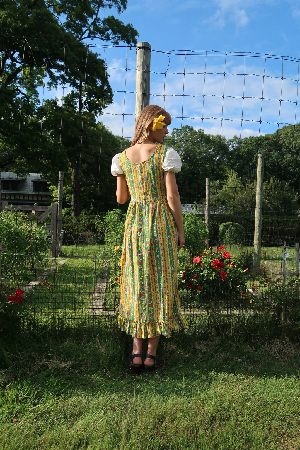 A woman stands outdoors in a garden wearing a vintage midi dress with a yellow, green, and orange floral pattern, white puff sleeves, and a back zipper. She is viewed from behind, looking out towards trees and a wire fence.