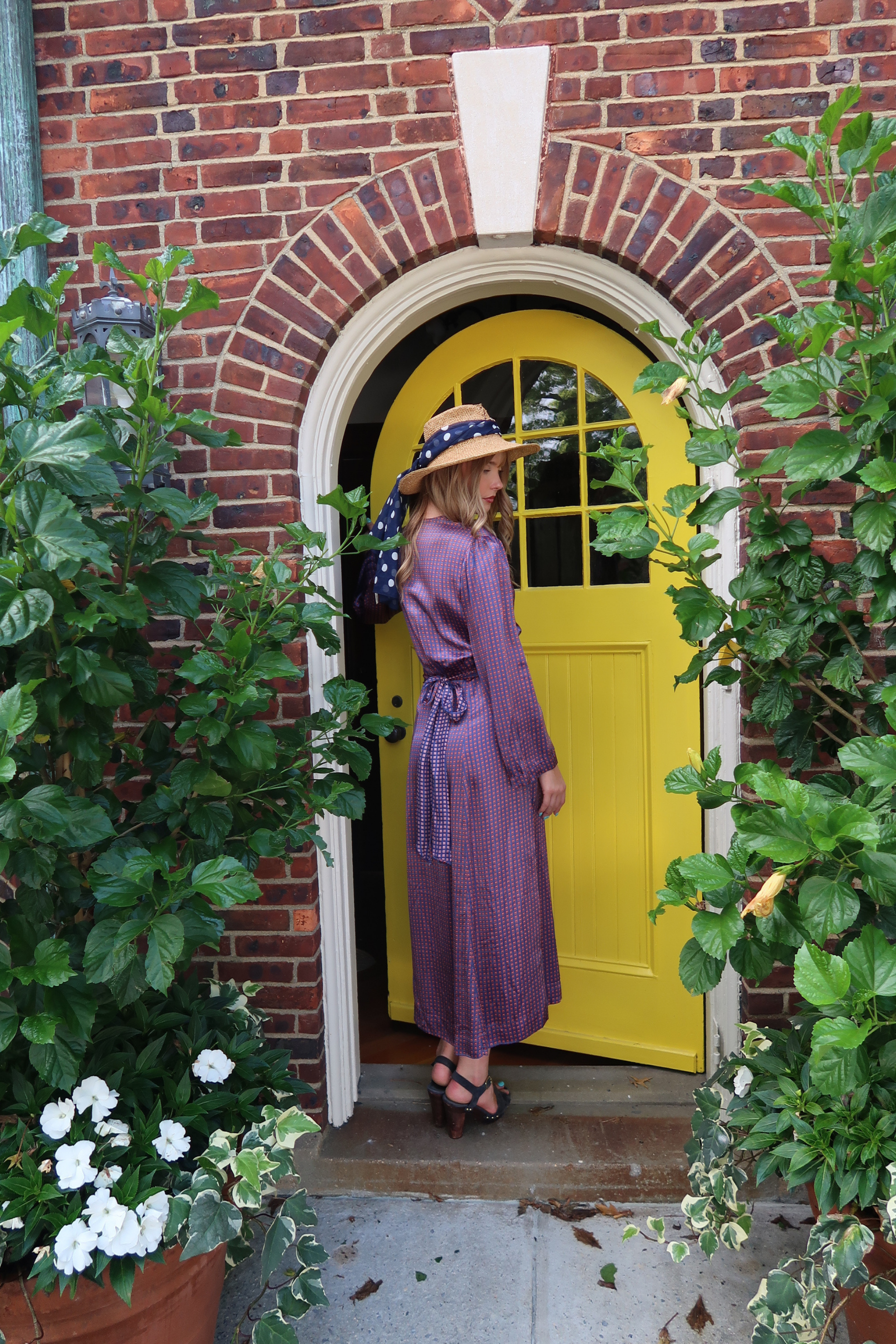 Back view  of model wearing a vintage silk dress standing in front of an open bright yellow front door set within a red brick archway, framed by green plants.